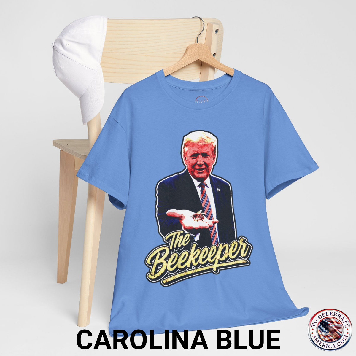 Carolina Blue T‑shirt featuring a pop‑art grunge illustration of President Trump holding a bee in his hand with bold ‘The Beekeeper’ text below.